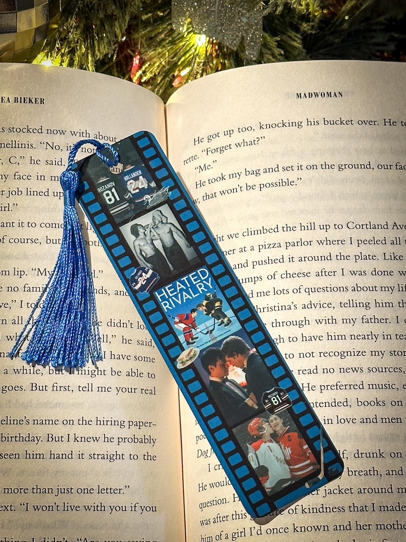 May include: A blue bookmark with a film strip design featuring hockey-themed images and the text "HEATED RIVALRY." The bookmark has a blue tassel and is resting on the pages of a book.