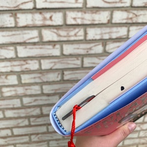 May include: A hardcover book with a red cover and a blue spine. A red tassel bookmark is inserted into the book. The book is held against a brick wall.