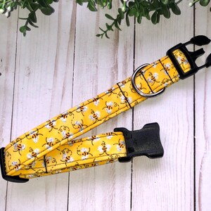 May include: A yellow dog collar with a bee pattern and black buckle. The collar is made of fabric and has a silver D-ring.