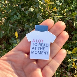 Puede incluir: Marcador de libro en forma de camiseta blanca con el texto "A LOT TO READ AT THE MOMENT" en letras negras y rosas.