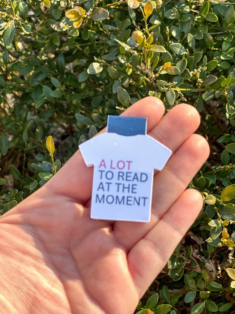 Puede incluir: Marcador de libro en forma de camiseta blanca con el texto "A LOT TO READ AT THE MOMENT" en letras negras y rosas.