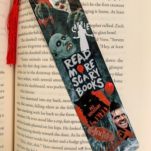 May include: A bookmark featuring a collage of horror movie characters and imagery. The bookmark is black and red with the text "Read More Scary Books" in white. The characters include Pennywise the clown from "IT", a creature from "Carrie", and a character from "Pet Sematary".