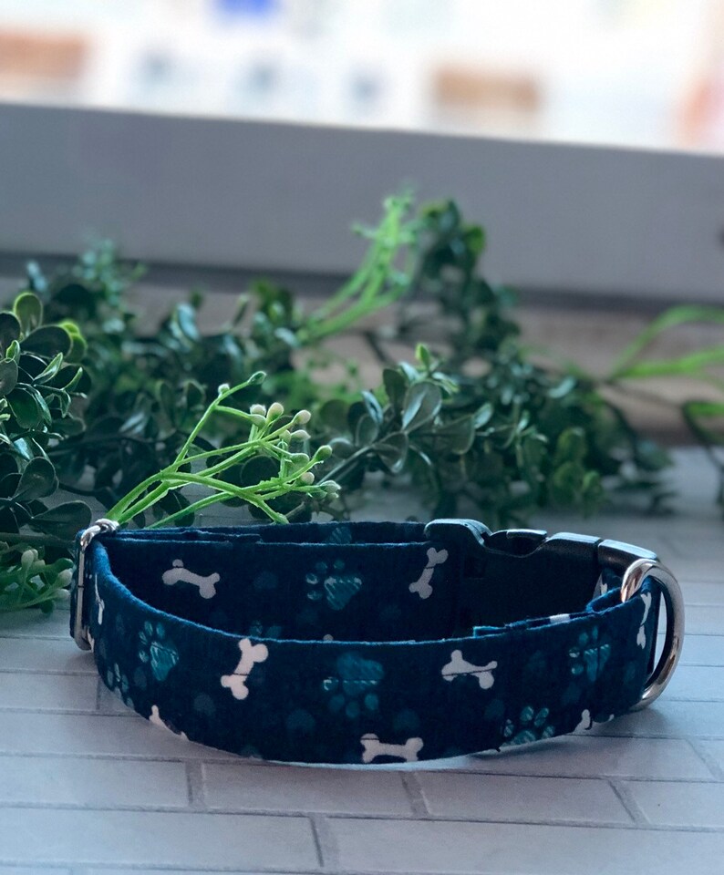 Puede incluir: Un collar para perro azul oscuro con un patr&oacute;n de huesos y huellas blancas. El collar tiene una hebilla plateada y un anillo en D plateado.