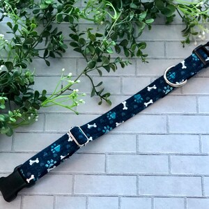 Puede incluir: Un collar de perro azul con huellas blancas y dise&ntilde;os de huesos. El collar tiene una hebilla plateada y un anillo en D plateado.