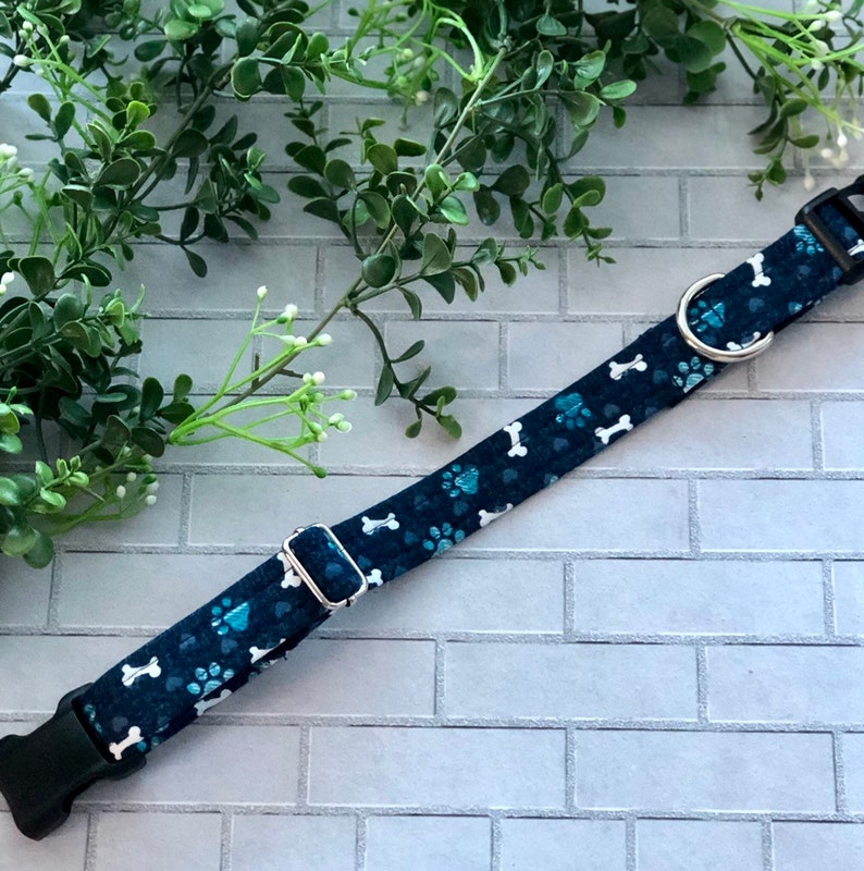 Puede incluir: Un collar de perro azul con huellas blancas y dise&ntilde;os de huesos. El collar tiene una hebilla plateada y un anillo en D plateado.