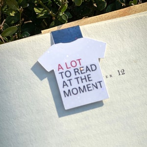 Puede incluir: Un marcap&aacute;ginas con forma de camiseta blanca con el texto "A LOT TO READ AT THE MOMENT" en letras rojas y negras.