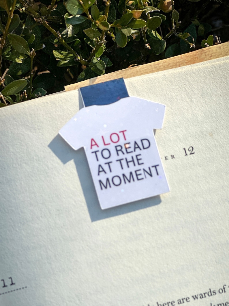 Puede incluir: Un marcap&aacute;ginas con forma de camiseta blanca con el texto "A LOT TO READ AT THE MOMENT" en letras rojas y negras.