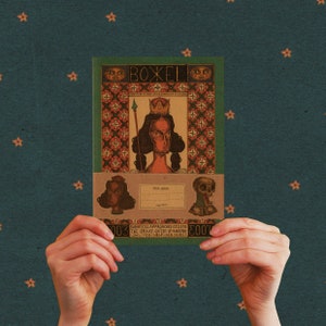 Puede incluir: Una cubierta de cuaderno de estilo vintage con una ilustración colorida de una reina con una corona y una lanza. La cubierta tiene un patrón geométrico y el texto "BOXEL" en la parte superior. El texto "YOUR NAME" está impreso en una etiqueta blanca debajo de la ilustración. El texto "DARKNESS: APPROACHES GYLLENE THE BRAVE QUEEN OF HAARPLA CAN YOU HELP HER SHIMEN" está impreso en una etiqueta negra en la parte inferior.