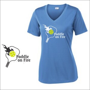 May include: A light blue v-neck t-shirt with a white graphic of a paddle and ball on fire. The text "Paddle on Fire" is printed below the graphic.