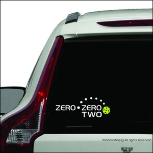 May include: A white car with a black rear window. The rear window has a white vinyl decal that says "ZERO ZERO TWO" with a yellow pickleball in between the words.