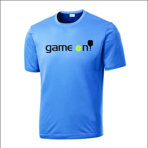 May include: Light blue t-shirt with the words "game on!" in black, a yellow pickleball, and a black pickleball paddle graphic. The shirt is short-sleeved and made of a smooth fabric.