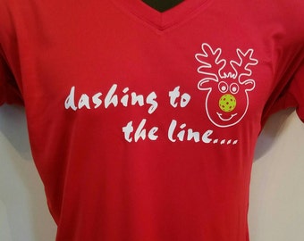 May include: Red v-neck t-shirt with a white design of a reindeer with a yellow and green pickleball for a nose. The text "dashing to the line..." is written below the reindeer.