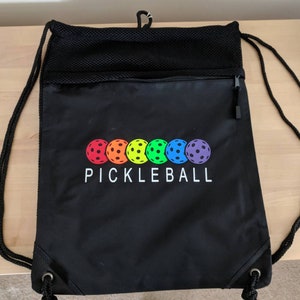 May include: Black drawstring bag with a mesh pocket and a zippered pocket. The bag has a rainbow-colored pickleball logo with the word "PICKLEBALL" printed below it.