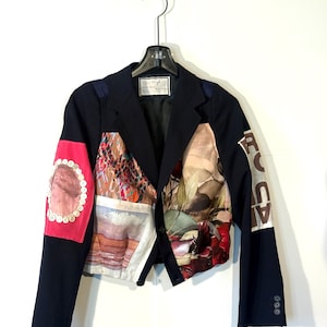 May include: A navy blue blazer with a patchwork design. The blazer features a variety of patterns and colors, including floral, abstract, and portrait prints. The sleeves have a three-button closure. The blazer is hanging on a black hanger.