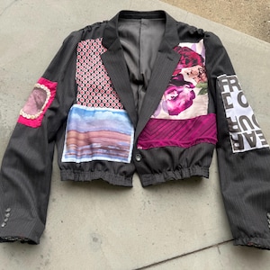 Puede incluir: Una chaqueta corta de color gris carbón con rayas diplomáticas que presenta varios parches de tela, incluyendo un estampado floral, una escena de paisaje, un cuadrado rosa con botones blancos y un parche blanco con el texto negro "FREEDOM" y "EAR".
