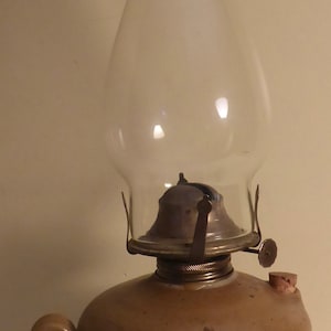 May include: A brown ceramic oil lamp with a floral design and a clear glass chimney. The lamp has a metal burner and a cork stopper.