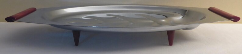 May include: A silver serving tray with a raised rim and three red legs. The tray has a slightly oval shape and a smooth surface.