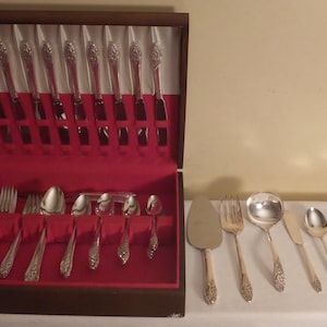 May include: A silver-colored flatware set with ornate floral detailing, displayed in a wooden case with a red velvet interior. Additional pieces are arranged on a white surface, including a cake server, forks, spoons, and knives.