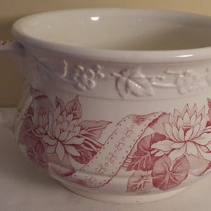 Può includere: Un vaso da notte in ceramica bianca con un motivo floreale rosa. Il vaso ha un manico e un'ampia apertura rotonda.