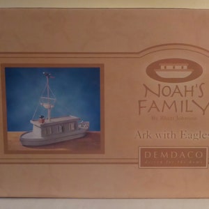 Può includere: Una scatola marrone con un'immagine bianca di un'arca di legno con due aquile in cima. La scatola ha il testo "Noah's Family" e "Ark with Eagles". La scatola è della società "Demdaco".