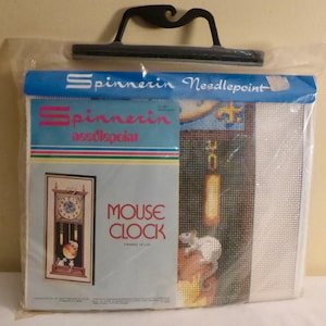 May include: A needlepoint kit featuring a clock with a mouse on it. The kit is packaged in a clear plastic bag with a blue header that reads "Spinnerin Needlepoint" and "Mouse Clock". The finished size is 10" x 27".