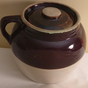 May include: A brown and white glazed ceramic crock with a lid. The crock has a single handle and a brown glaze on the top half and a white glaze on the bottom half.