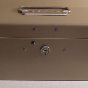 May include: A beige metal lock box with a silver handle and a keyhole. The box has a slightly textured surface.
