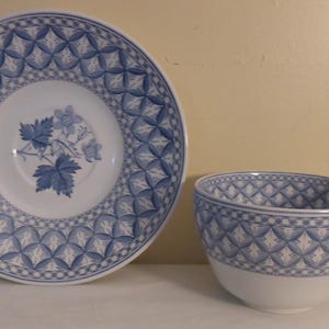 May include: A white teacup and saucer set with a blue floral and geometric pattern. The saucer features a central floral design, while the cup and saucer have a repeating diamond pattern. The set is ideal for serving tea.