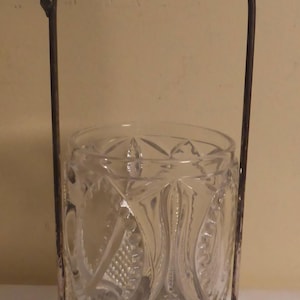 May include: A clear glass jar with a decorative etched pattern, held within a silver-colored metal frame. The frame has an arched handle with ornate detailing at the top and base. The jar is cylindrical.