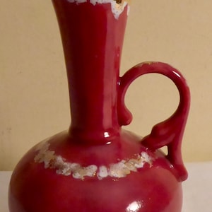May include: A red ceramic jug with a white and gold rim and a handle. The jug has a rounded body and a long, thin neck.