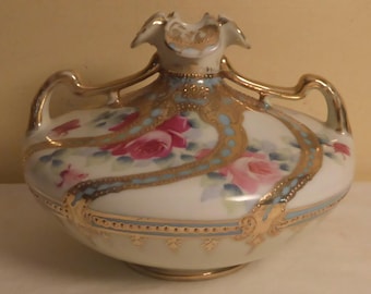 May include: A white porcelain vase with pink roses and gold detailing. The vase features two gold handles and a ruffled top. Blue and gold decorative bands encircle the vase. The floral design is hand-painted.