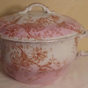 May include: A white ceramic chamber pot with a pink and brown floral design. The pot has a lid with a handle.