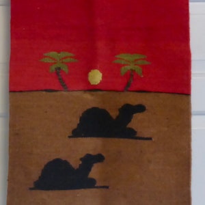 May include: A rectangular wall hanging with a desert scene. The top portion is red, with two green palm trees and a yellow sun. The bottom portion is brown with two black camel silhouettes. White fringe hangs from the bottom.