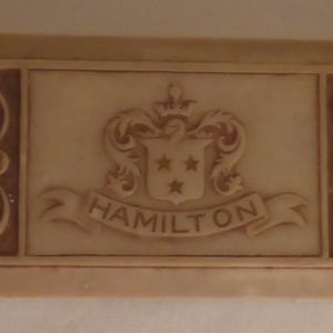 May include: A rectangular wooden box with a carved design. The box is light brown and features a crest with the word "HAMILTON" written on a banner.