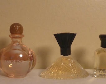 May include: Five perfume bottles of varying shapes and sizes are displayed. The bottles are filled with amber-colored liquid and have different cap designs. The bottles are arranged on a white surface against a neutral background.