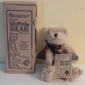 May include: A beige mohair bear wearing a red and black plaid scarf. The bear is sitting on a white surface and is holding a small tag that reads "The Boyds Collection Original Mohair Bear Collection Adam F. Bearington Guaranteed Bears & Hares You Can Trust..."