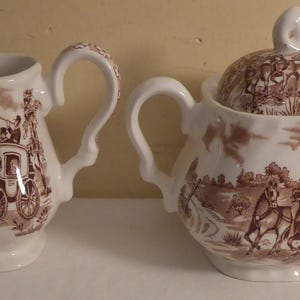 May include: A white porcelain creamer and sugar bowl set with brown floral designs and a scene of a horse-drawn carriage. The creamer has a handle and a spout, while the sugar bowl has a lid.