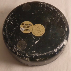 Pu&ograve; includere: Un oggetto rotondo in pietra ollare nera con venature bianche. Il fondo presenta tre piccoli piedini rotondi e due adesivi. Un adesivo recita "GENUINE SOAP STONE", e l'altro dice "HANDCRAFTED IN ORIGINAL STONE MADE IN CANADA".