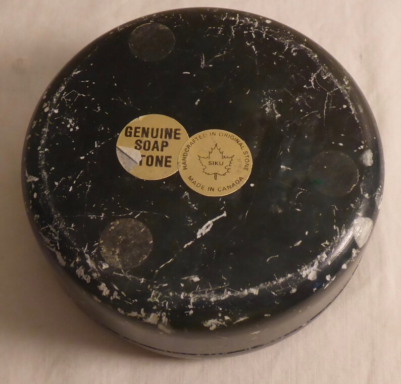 Pu&ograve; includere: Un oggetto rotondo in pietra ollare nera con venature bianche. Il fondo presenta tre piccoli piedini rotondi e due adesivi. Un adesivo recita "GENUINE SOAP STONE", e l'altro dice "HANDCRAFTED IN ORIGINAL STONE MADE IN CANADA".