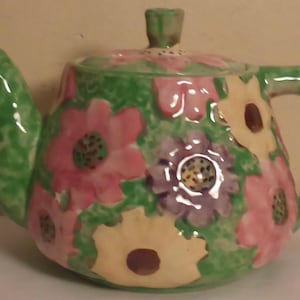 May include: A green, pink and white floral teapot and creamer set. The teapot has a lid and a handle. The creamer has a handle. The set is made of ceramic.