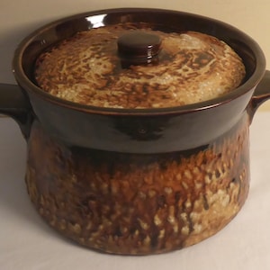 May include: A brown and beige ceramic casserole dish with a lid. The dish has a mottled glaze and two handles.
