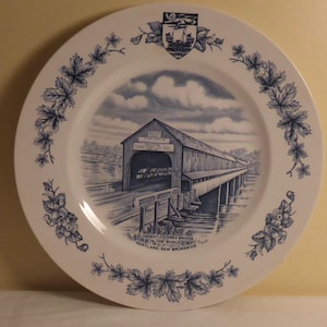 May include: A white ceramic plate with a blue and white design of a covered bridge. The plate has a blue border with a floral design. The text on the plate reads "Longest Covered Bridge in the World, Hartland, New Brunswick".