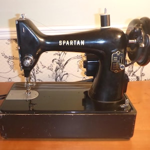 May include: A vintage black Spartan sewing machine. The machine has a black base and body with the word "SPARTAN" in white lettering. The machine has a hand wheel, needle, and thread spool. The machine is on a wooden surface.