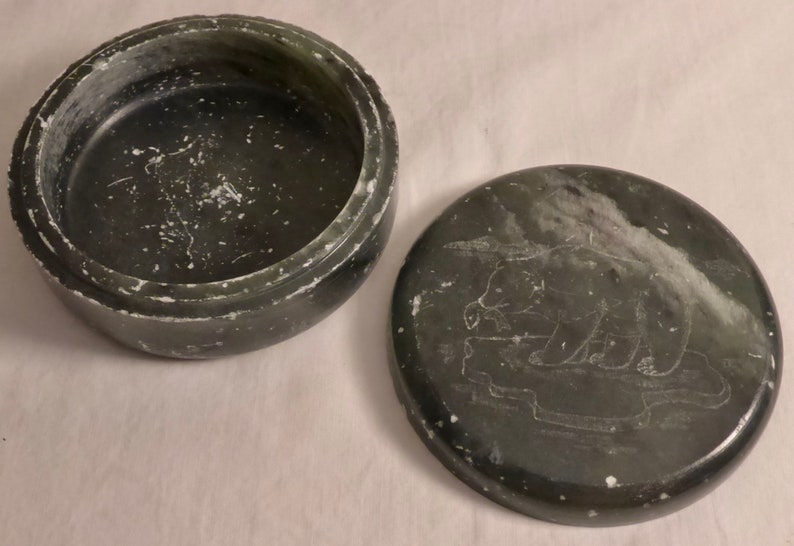 Pu&ograve; includere: Scatola portagioie rotonda verde scuro con coperchio. Il coperchio presenta un disegno di orso inciso. La scatola &egrave; aperta, rivelando l'interno. La scatola e il coperchio hanno un aspetto strutturato con accenti bianchi.