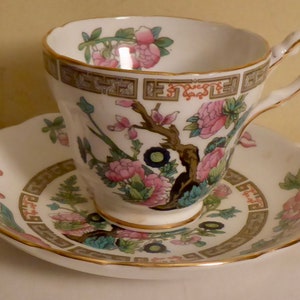 May include: A white teacup and saucer with a floral pattern. The pattern features pink and blue flowers, green leaves, and a brown tree branch. The cup and saucer have a gold trim.