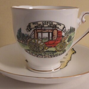 May include: A white teacup and saucer with a gold rim. The teacup features a colourful illustration of a red stagecoach with the text "Red Coach Inn 100 Mile House British Columbia".