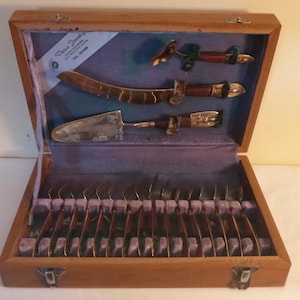 May include: A vintage wooden cutlery set with a velvet-lined interior. The set includes a cake server, a curved knife, and other utensils with ornate handles. The box is closed with a metal clasp. The set is from Venus Jewelry.