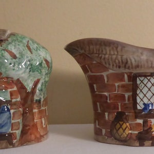 May include: A brown and white ceramic sugar bowl and creamer set with a cottage scene. The sugar bowl is shaped like a cottage with a thatched roof and a window. The creamer is shaped like a pitcher with a handle and a window. Both pieces feature a brick wall, a tree, and a person looking out the window.