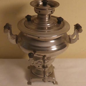 May include: A silver samovar with a black handle and a black knob on the spout. The samovar has a round body and a tall, cylindrical spout. It sits on a square base with four legs.