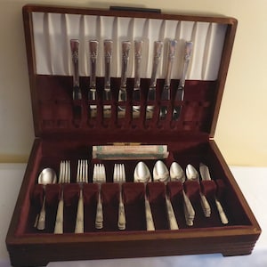 May include: A wooden box with a burgundy velvet interior containing a set of silver cutlery. The cutlery includes forks, knives, and spoons. The box has a label that reads "WARRANTY".